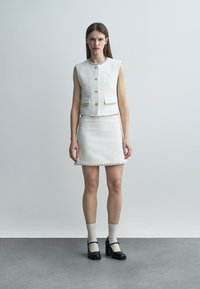 Naine seisab, kandes valget tekstuuriga varrukateta jopet ja sobivat seelikut, valged sokid ja mustad Mary Jane stiilis kontsakingad, lihtsa tausta ees.