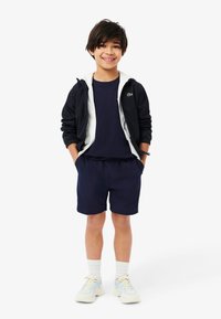 Navy shorts and T-shirt paired with a black and white zip-up jacket. Light sneakers with pastel accents. Model stands with hands in pockets.