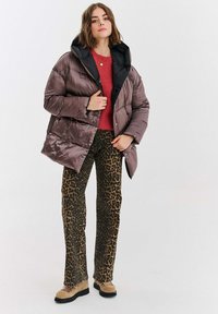 Brązowa kurtka puchowa z czarnym kapturem, noszona na czerwonym swetrze, zestawiona z spodniami w panterkę i beżowym obuwiem z czarnymi podeszwami.