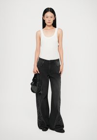 Black wide-leg jeans with side branding, textured fabric; paired with a white tank top and a mesh bag. Model stands with hands on hips.