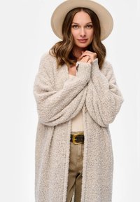 Długi kardigan z otwartym przodem wykonany z teksturowanego, jasnobeżowego dzianinowego materiału. Modelka ma na sobie kremowy top i beżowe spodnie, do tego szerokim rondem jasny kapelusz.