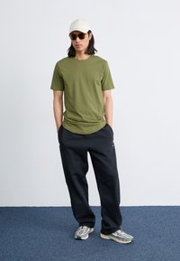 Olivgrünes Baumwoll-T-Shirt, schwarze Trainingshose, cremefarbene Mütze, dunkle Sonnenbrille und graue Sneakers mit hellen Akzenten. Schlichtes Design und lässige Passform.