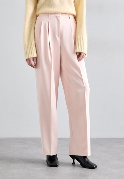 Pantalones de tiro alto en rosa claro, con pliegues y corte entallado, combinados con un suéter de punto amarillo y tacones negros de punta. Textura de tela suave.