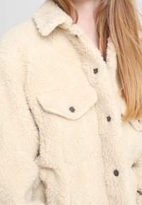 Veste en shearling crème floue avec un col, fermeture à bouton-pression et deux poches poitrines avec des boutons foncés.