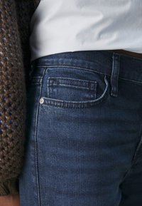 Jeans in denim blu scuro con una vestibilità slim, dotati di una tasca standard e dettagli di cucitura chiara. Indossati con un maglione lavorato a maglia marrone texturizzato.