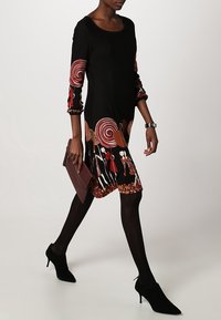 Femme marchant, portant une robe noire avec des motifs en spirale rouge et blancs, des collants noirs, des talons noirs, et tenant une pochette bordeaux.