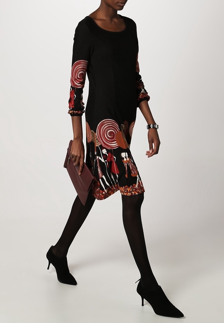 Femme marchant, portant une robe noire avec des motifs en spirale rouge et blancs, des collants noirs, des talons noirs, et tenant une pochette bordeaux.