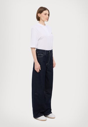 White short-sleeve polo shirt paired with dark blue wide-leg denim jeans. Notable details include stitching and a relaxed fit, worn with white shoes.