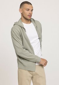 Haut à capuche zippé vert clair avec un cordon de serrage et des poches avant, superposé sur un t-shirt blanc. Texture douce et coupe décontractée.