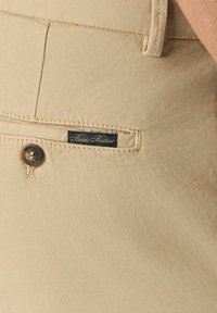 Beżowa bawełniana tkanina o gładkiej fakturze, z zapięciem na guzik i małą, granatową metką, na której widnieje złoty napis "Brooks Brothers".