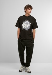 Sort oversized t-shirt med hvid grafisk tryk, parret med sorte joggingbukser og metalliske sneakers, stående mod en neutral baggrund.