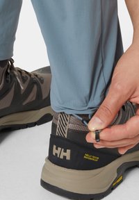 Vaaleansiniset ulkohousut säädettävillä lahkeensuilla, yhdistettynä vedenpitäviin tummanruskeisiin vaelluskengiin, joissa on teksturoitu takaosa ja logoyksityiskohta.