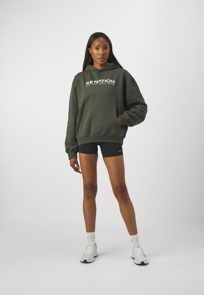 Felpa verde oliva con tasca frontale e testo del logo, abbinata a pantaloni corti neri e sneakers bianche, su uno sfondo chiaro.