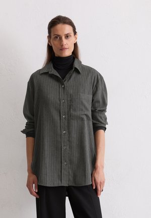 Camicia a righe grigie con colletto, dotata di taschino frontale e maniche arrotolate, abbinata a un top nero a collo alto.