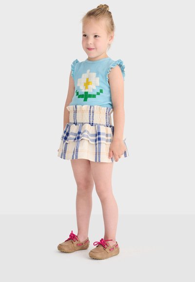 Jeune fille aux cheveux blonds portant un haut bleu sans manches avec une fleur blanche, une jupe à volants à carreaux et des chaussures marron avec des lacets roses, debout et souriante.