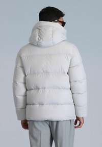 Veste matelassée gris clair avec capuche, présentant un design en patchwork, une texture douce et des poignets élastiques. Style minimaliste sans logo visible.