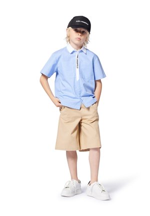 Hellblaues Kurzarmhemd mit vertikalen Streifen und weißem Kragen; beigefarbene Shorts; schwarze Kappe mit Logo; weiße Sneaker.