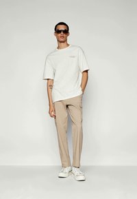 Jongeman in een wit oversized t-shirt, beige cargo pants, witte sneakers en zwarte zonnebril, staand met één hand in zijn zak.