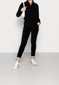 Salopette en velours côtelé noir avec un devant à boutons, deux poches poitrine et des jambes ajustées, associée à des baskets blanches avec des accents noirs.