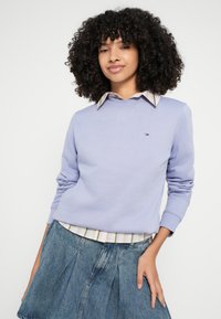 Gaiši violets džemperis ar apaļu kakla izgriezumu un garām piedurknēm, kārtots virs svītrainas kreklas ar apkakli, kopā ar džinsa svārkiem.