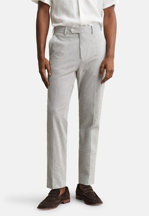 Άνδρας που φοράει ανοιχτό γκρι υφασμάτινο παντελόνι, λευκό κοντομάνικο πουκάμισο κεντημένο μέσα και σκούρα καφέ σουέντ μοκασίνια, στέκεται μπροστά σε λευκό φόντο.