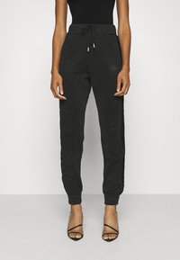 Schwarze Sweatpants mit glitzernder Textur, ausgestattet mit einem Bindegürtel, seitlichen Streifen und engen Bündchen, kombiniert mit schwarzen Riemchenheels.