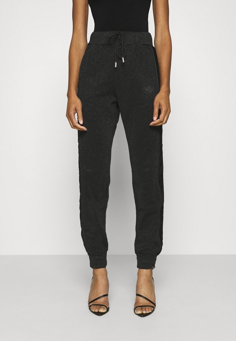 Schwarze Sweatpants mit glitzernder Textur, ausgestattet mit einem Bindegürtel, seitlichen Streifen und engen Bündchen, kombiniert mit schwarzen Riemchenheels.