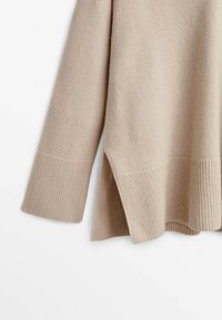 Manche et ourlet d'un pull en maille beige avec larges poignets côtelés et détail fendu sur le côté, sur fond blanc uni.