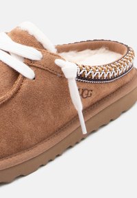 Pruun seemisnahast mokassiin valgete paeltega, pehme fliisvoodriga, kaela ümber dekoratiivse õmblusega ja tekstureeritud kummist tallaga.