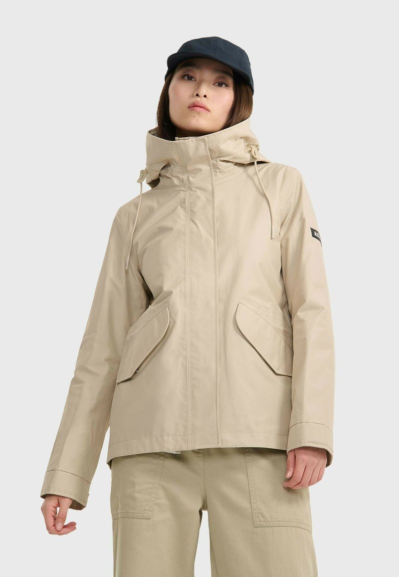 Veste imperméable beige avec capuche, fermeture éclair à l'avant et grandes poches latérales. Dispose de cordons de serrage réglables au niveau du cou. Texture lisse.