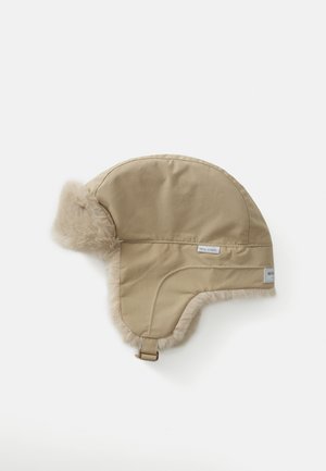 Chapeau beige en fourrure avec une forme arrondie, un matériau extérieur épais et une sangle sur le côté pour un maintien sécurisé. Présente un détail d'étiquette de marque.