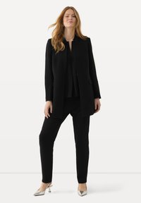 Zwarte op maat gemaakte pak met een lange jas en aangesloten broek, met een gladde textuur, en gecombineerd met metallic zilveren hakken.