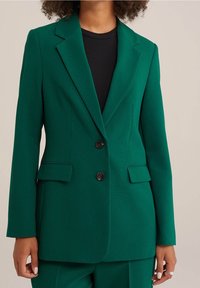 Grön kavaj i stretchtyg, med slagkrage, två framfickor och två svarta knappar, med en slät yta.