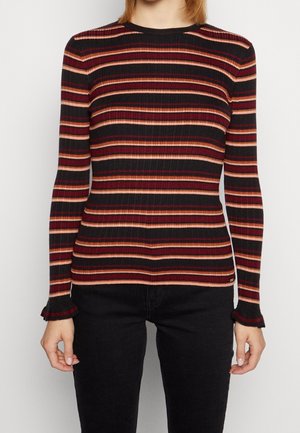 Femme portant un haut côtelé à manches longues avec des rayures horizontales rouges, roses et noires, associé à un pantalon noir sur un fond uni.