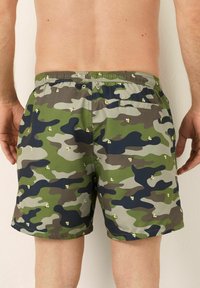 Costume da bagno mimetici con una combinazione di motivi verdi, grigi e neri, caratterizzati da piccoli fiori gialli e una vita elastica.