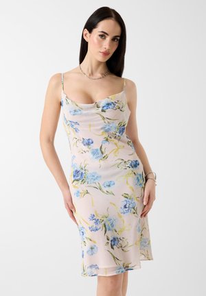 Donna con lunghi capelli scuri, che indossa un vestito senza maniche di colore rosa chiaro con una stampa floreale blu, posando contro uno sfondo bianco.