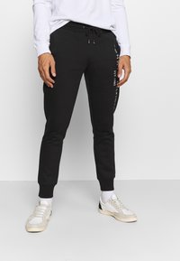 Zwarte joggingsbroek met een taperende pasvorm, gemaakt van katoen, een elastische tailleband met trekkoord en witte tekstaccenten aan de zijkant.