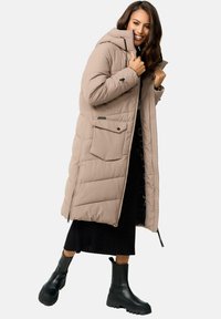 Long beige puffer coat with a hood, zip closure, two front pockets, and prominent stitching. Worn over a black ribbed skirt and black boots.