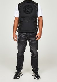 Veste matelassée noire avec logo en relief et sangles réglables, associée à un jean noir déchiré et des baskets noires et grises.