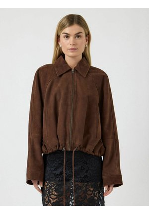 Ženska s ravnimi blond lasmi, oblečena v rjavo semiš usnjeno jakno z zadrgo in črno čipkasto krilo, stoji pred enobarvnim svetlim ozadjem.