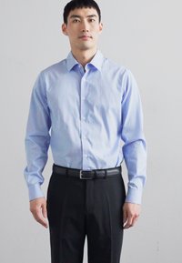 Camicia azzurra chiara e bianca a righe con bottoni, realizzata in un tessuto dalla texture liscia. Indossata con pantaloni neri e una cintura.