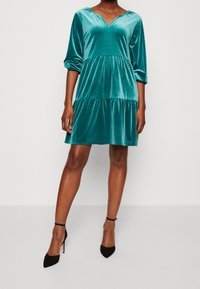 Tyrkysové sametové šaty s véčkovým výstřihem, tříčtvrtečními rukávy a sukni s volány. Mají měkkou texturu a volný, pohodlný střih.