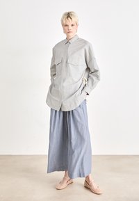 Camicia a righe grigia e bianca con bottoni; presenta due tasche frontali e dettagli con cordino. Abbinata a pantaloni larghi a righe blu chiaro e scarpe intrecciate.