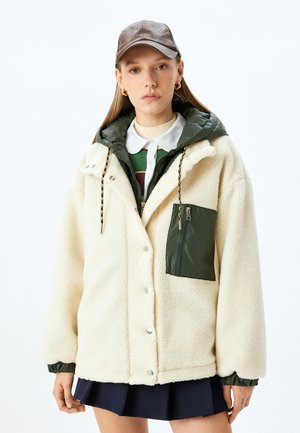 Veste en polaire de couleur crème avec des accents verts, poche zippée et capuche contrastante. Comprend des boutons pression et des détails avec cordon de serrage.