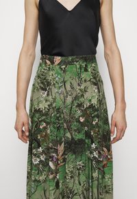 Femme portant un haut noir sans manches rentré dans une jupe verte avec un imprimé botanique détaillé comportant des feuilles et des fleurs.