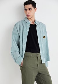 Chemise à rayures bleu-vert et blanche, associée à un t-shirt noir et un pantalon vert olive ; présente un patch sur la poche poitrine gauche.