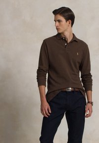 Brūns garām piedurknēm polo krekls ar apkakli, ar mazu logo uz krūšu daļas, apvienots ar tumši ziliem džinsiem. Gluda tekstūra.