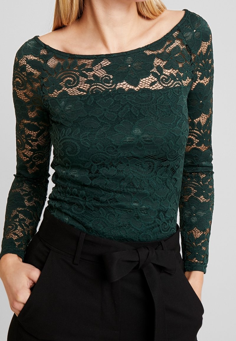 Femme portant un haut en dentelle florale vert foncé à manches longues, associé à un pantalon noir taille haute noué d’un nœud à la taille.
