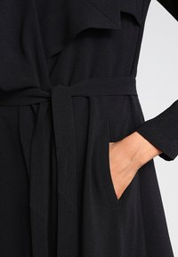 Rechte Hand in der Tasche eines schwarzen Mantels mit Gürtel und weitem Revers, zeigt Struktur und Stoffdetails.