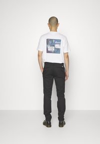 Un uomo con testa rasata sta in piedi di spalle, indossando una maglietta grafica bianca Adidas, pantaloni neri e scarpe nere su uno sfondo bianco uniforme.